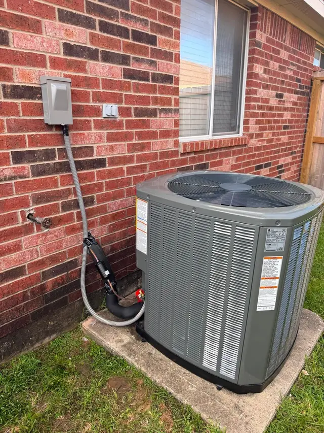 Outdoor condensing unit on a concrete pad against red brick, with its disconnect box and insulated line set